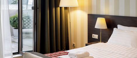 Ein Hotelzimmer mit Bett, zwei Nachttischlampen, einem Fenster mit Vorhängen und Blick ins Freie.