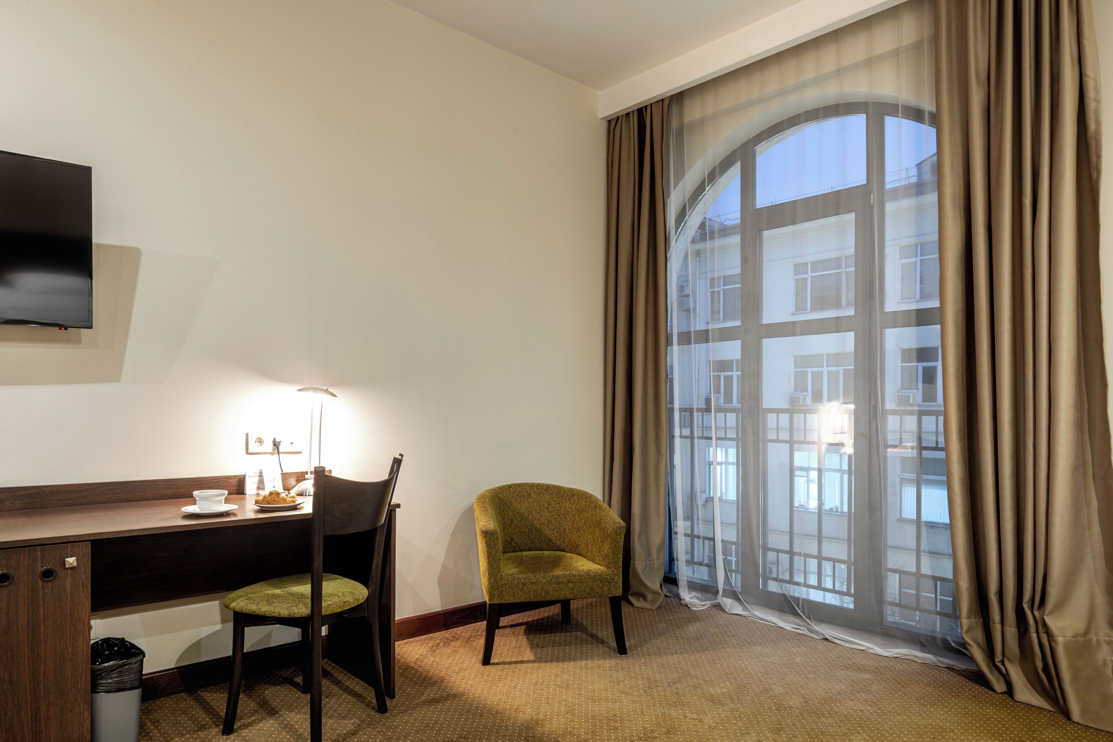 Habitación de hotel con escritorio, silla, televisor y un ventanal con cortinas.