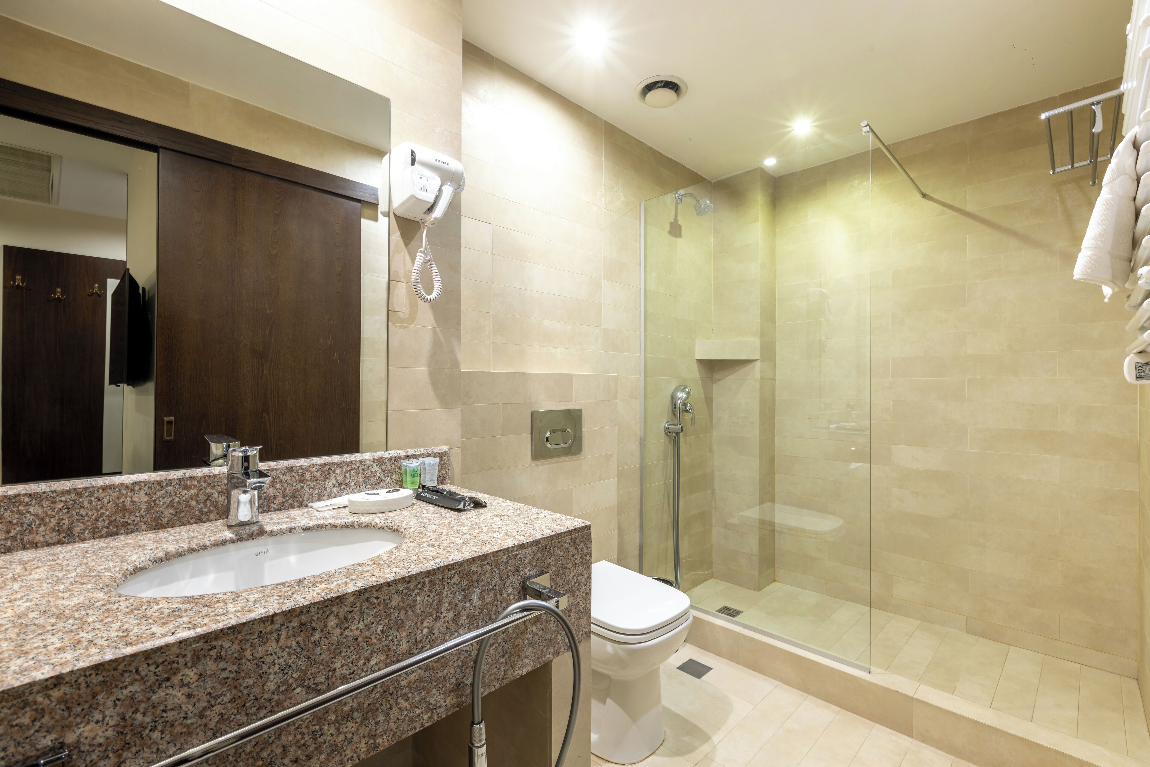 A bathroom with a granite countertop sink, a mirror, a hairdryer, and a glass-enclosed shower.
