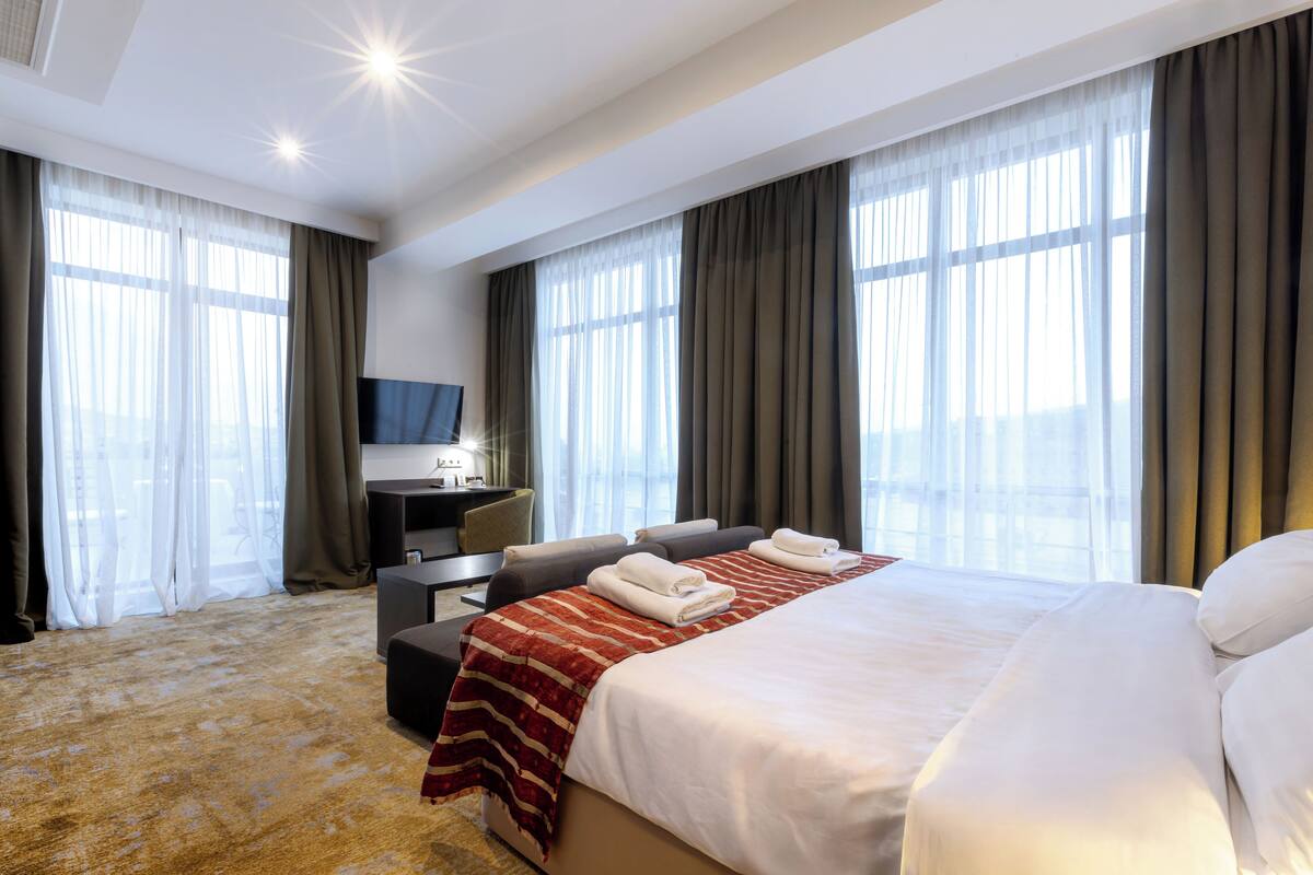 Habitación de hotel con una cama grande, un escritorio con silla, televisión y vistas a la ciudad a través de amplias ventanas.