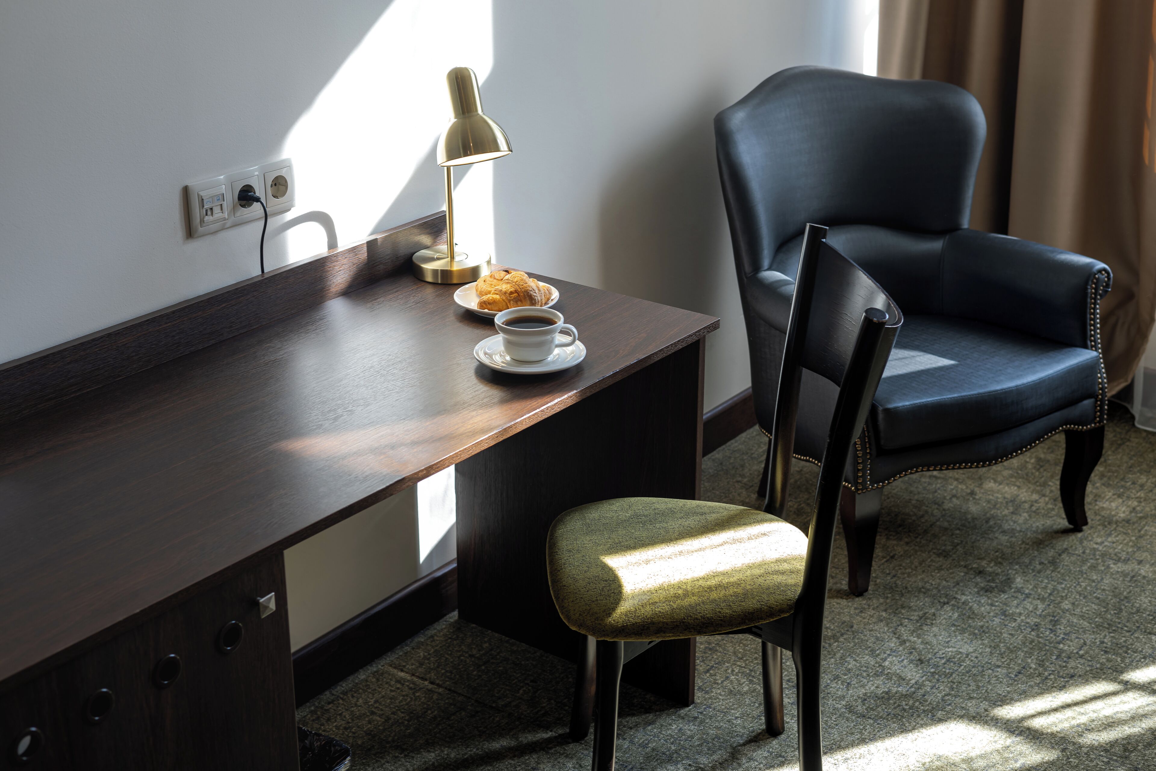 A desk with a lamp, a plate with a pastry, and a cup of coffee. There is a chair with a cushioned seat and backrest.
