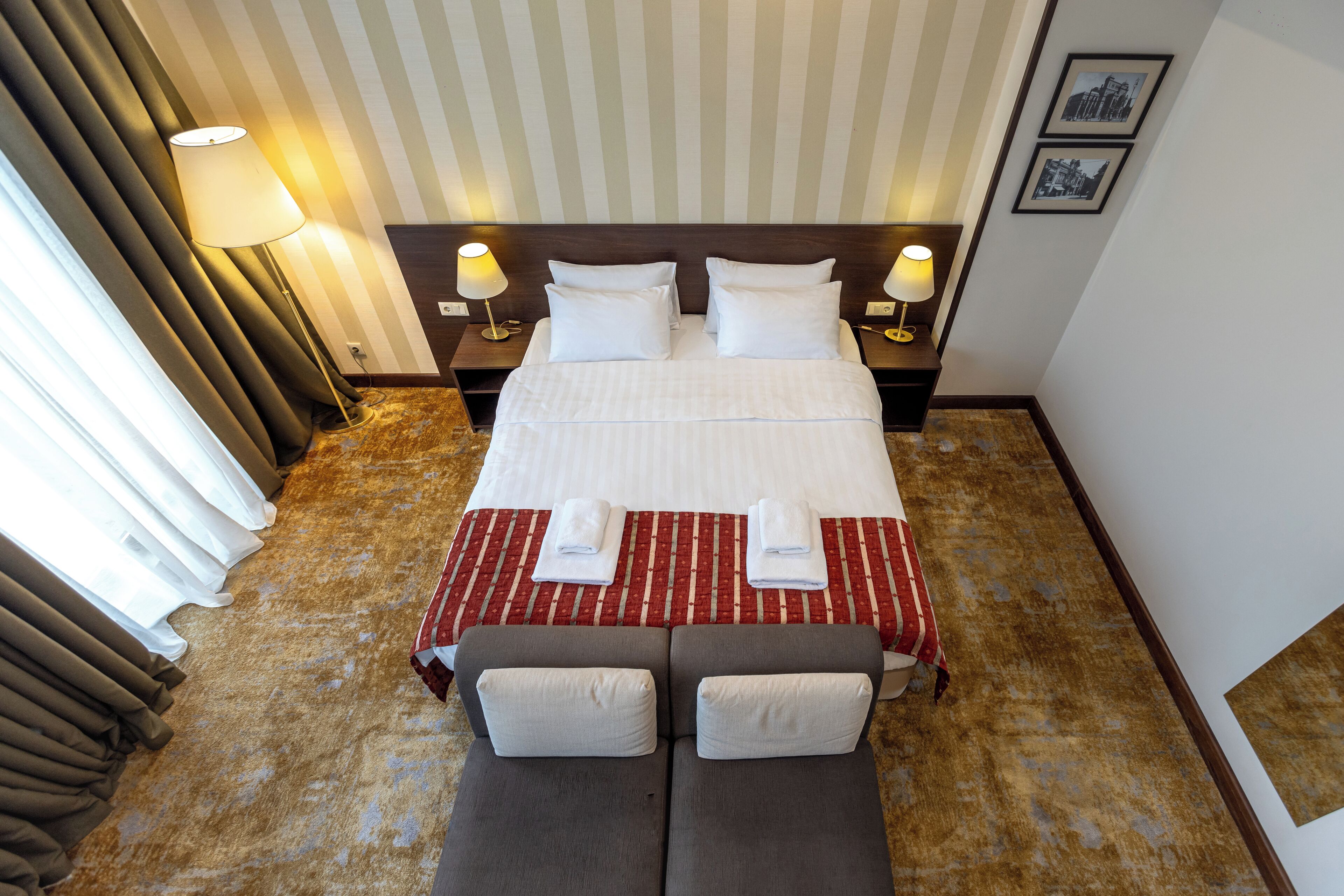 A hotel room with a large bed, two bedside lamps, a red and white striped bedspread, a grey sofa with pillows, and a carpeted floor.