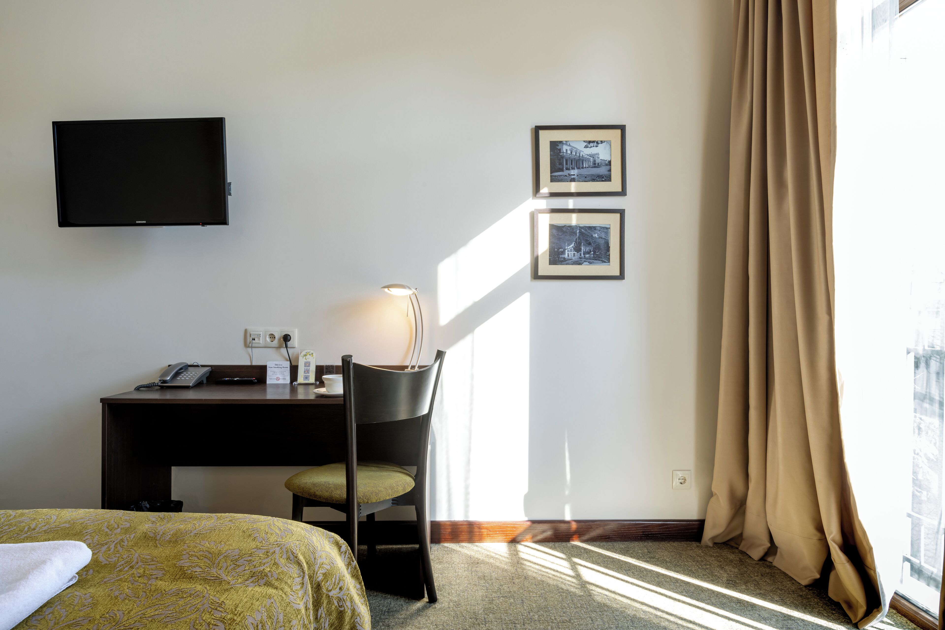 A hotel room with a bed, a desk with a lamp, a chair, and a TV mounted on the wall.