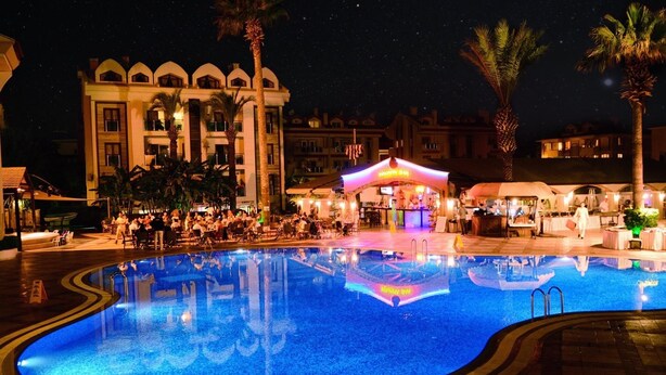 A nighttime swimming pool area with a building in the background, palm trees, and a brightly lit bar area. - Club Anastasia Hotel (Marmaris)