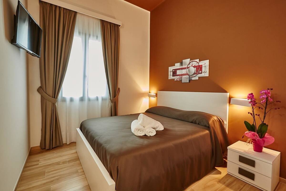 A neatly made bed with a brown bedspread and white pillows, a flat-screen TV mounted on the wall, a bedside table with a lamp and a vase of flowers, and a window with curtains.