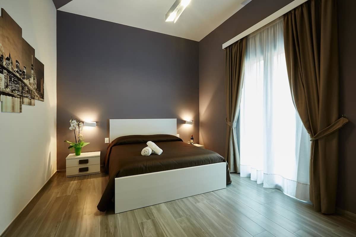 A modern bedroom with a large bed, a nightstand with a lamp, a potted plant, and a cityscape artwork on the wall.