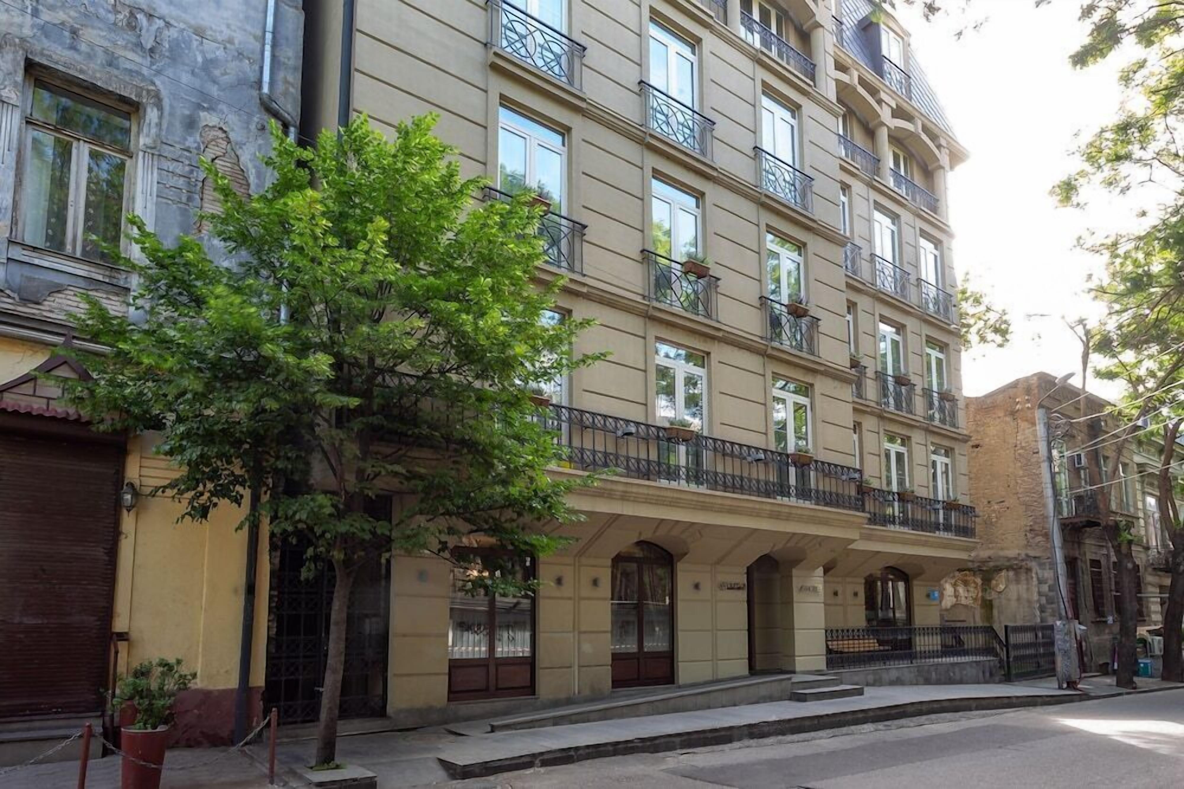 A multi-story building with balconies, large windows, and a tree in front.
