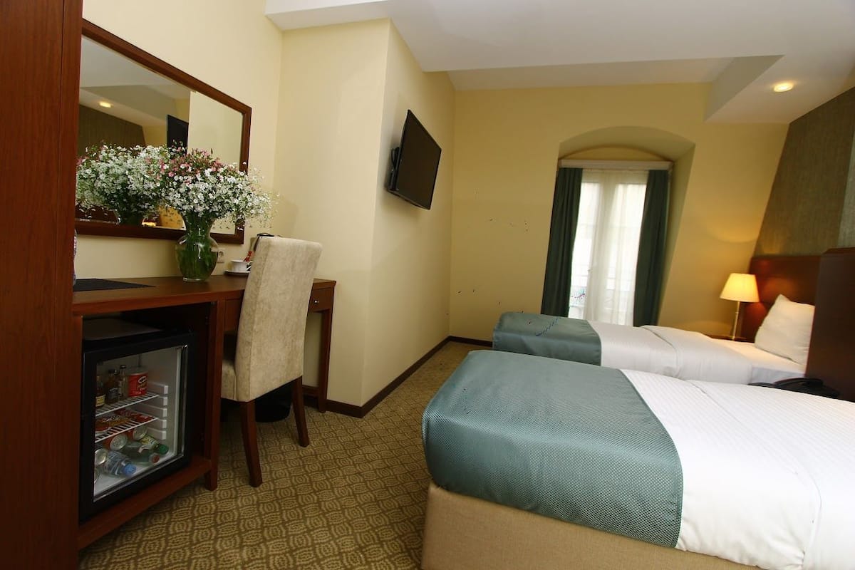 Une chambre d’hôtel avec deux lits, un bureau avec un miroir, une télévision, un réfrigérateur et une fenêtre avec des rideaux.