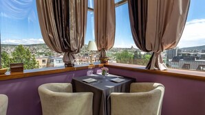 Un área de comedor con una mesa dispuesta para dos, ventanas amplias con cortinas y vistas a un paisaje urbano.