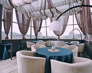 A modern dining area with round tables, comfortable chairs, and large windows offering a city view.