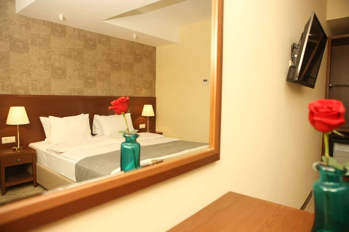 Une chambre d’hôtel avec un lit, deux tables de chevet avec des lampes, un téléviseur fixé au mur et un miroir qui reflète la pièce.