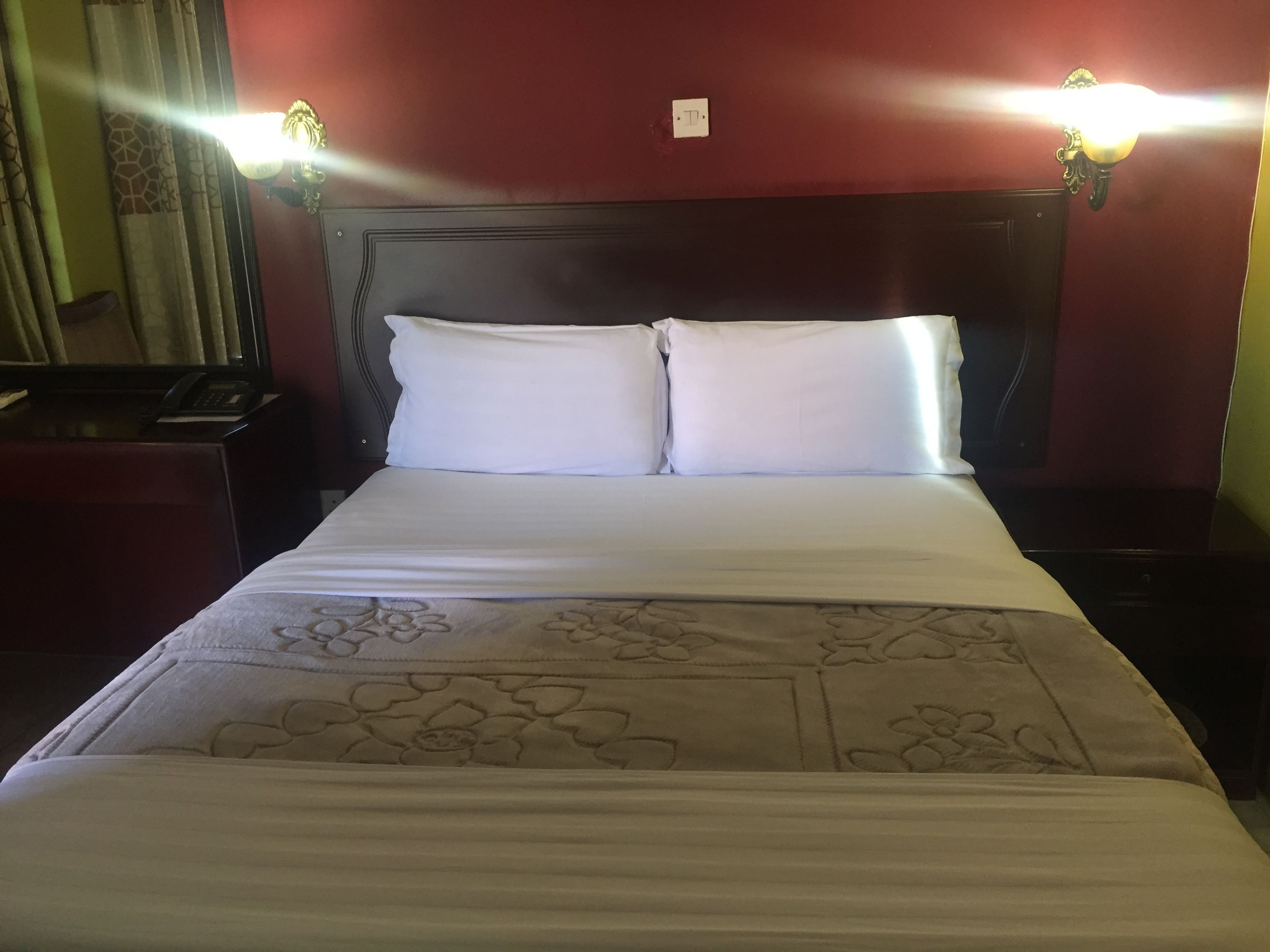 A neatly made bed with white linens and a floral-patterned bedspread. There are two bedside tables with lamps, and a telephone on one of the tables. The room has a dark headboard and red walls.