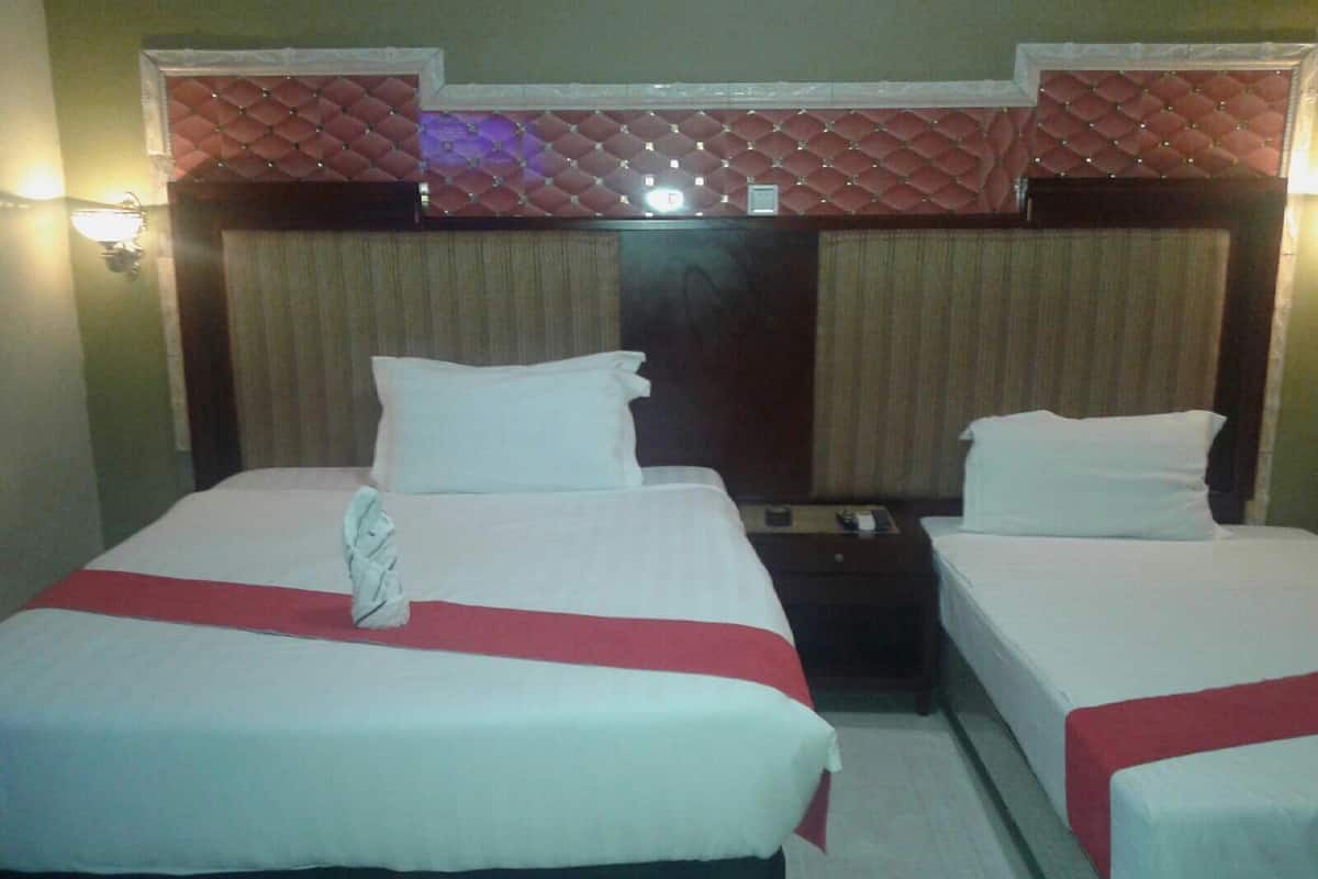 Habitación de hotel con dos camas, cada una con sábanas blancas y colchas rojas. Hay una mesita de noche entre las camas con una lámpara y una pequeña mesa. La habitación tiene un cabecero decorativo con un diseño de diamantes y un accesorio de iluminación encima.