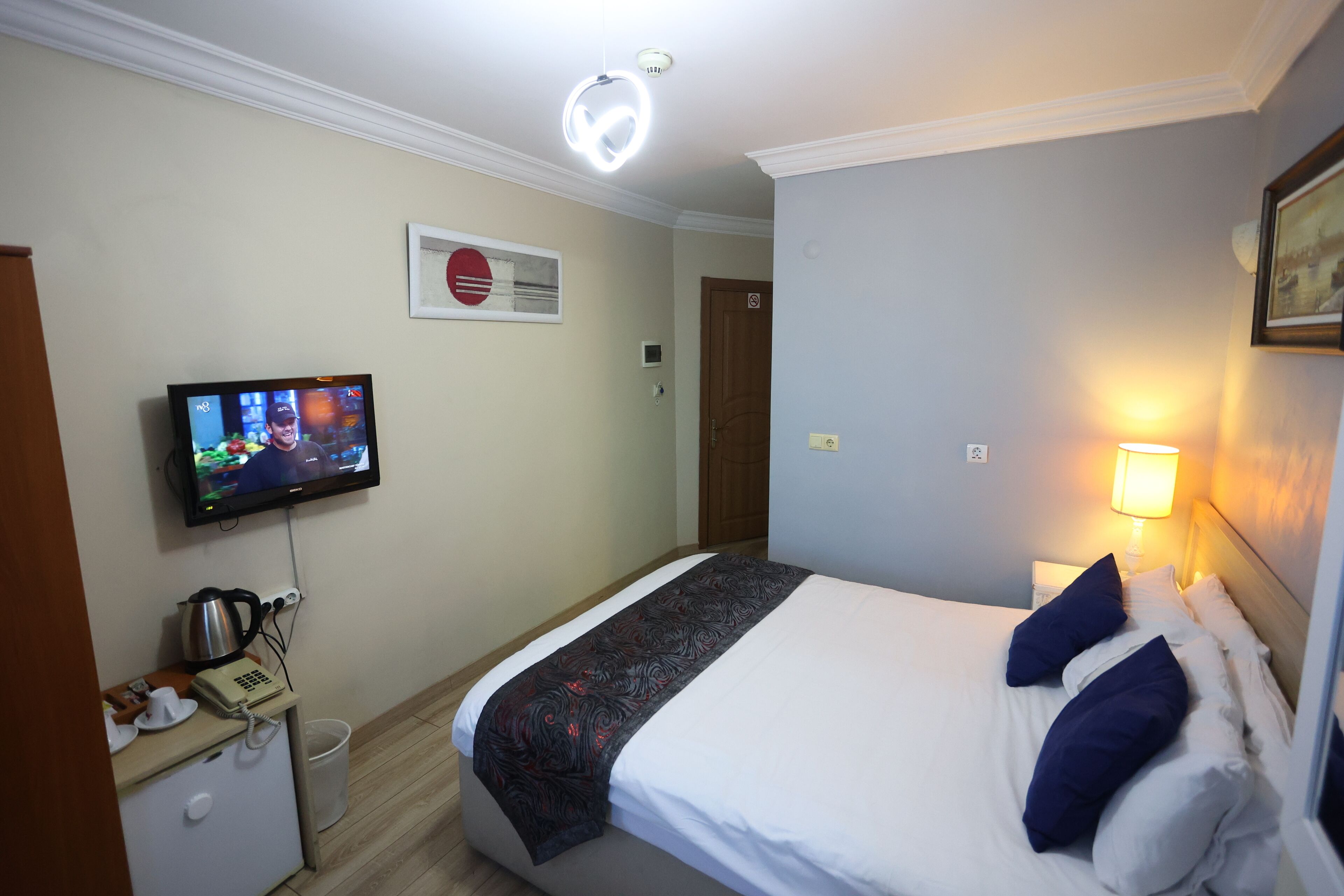 Ett hotellrum med en säng, en tv som är monterad på väggen, ett nattduksbord med en telefon och en lampa samt en dörr i bakgrunden.