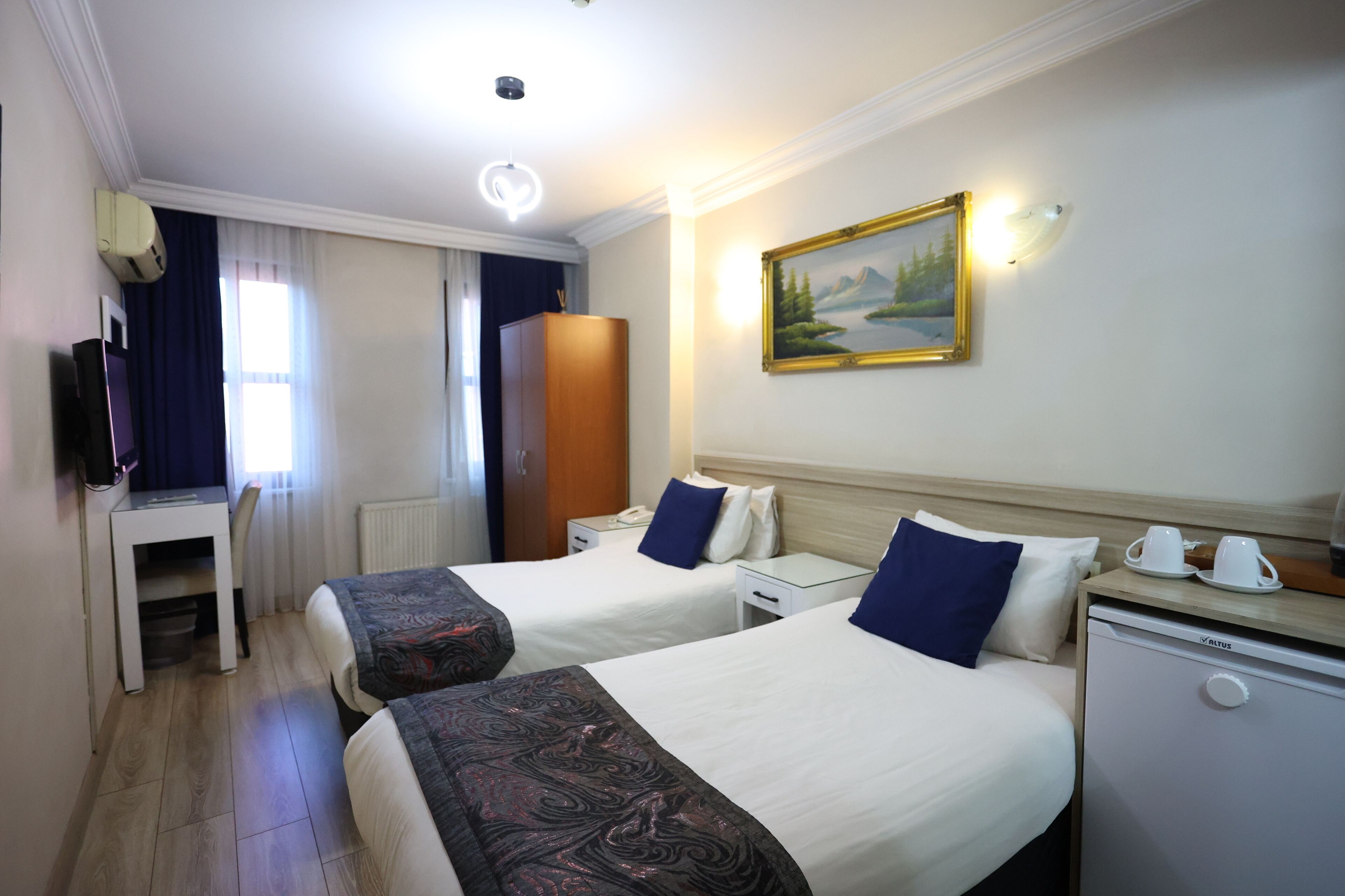 Quarto de hotel com duas camas, uma escrivaninha, um pequeno frigorífico e um quadro na parede.
