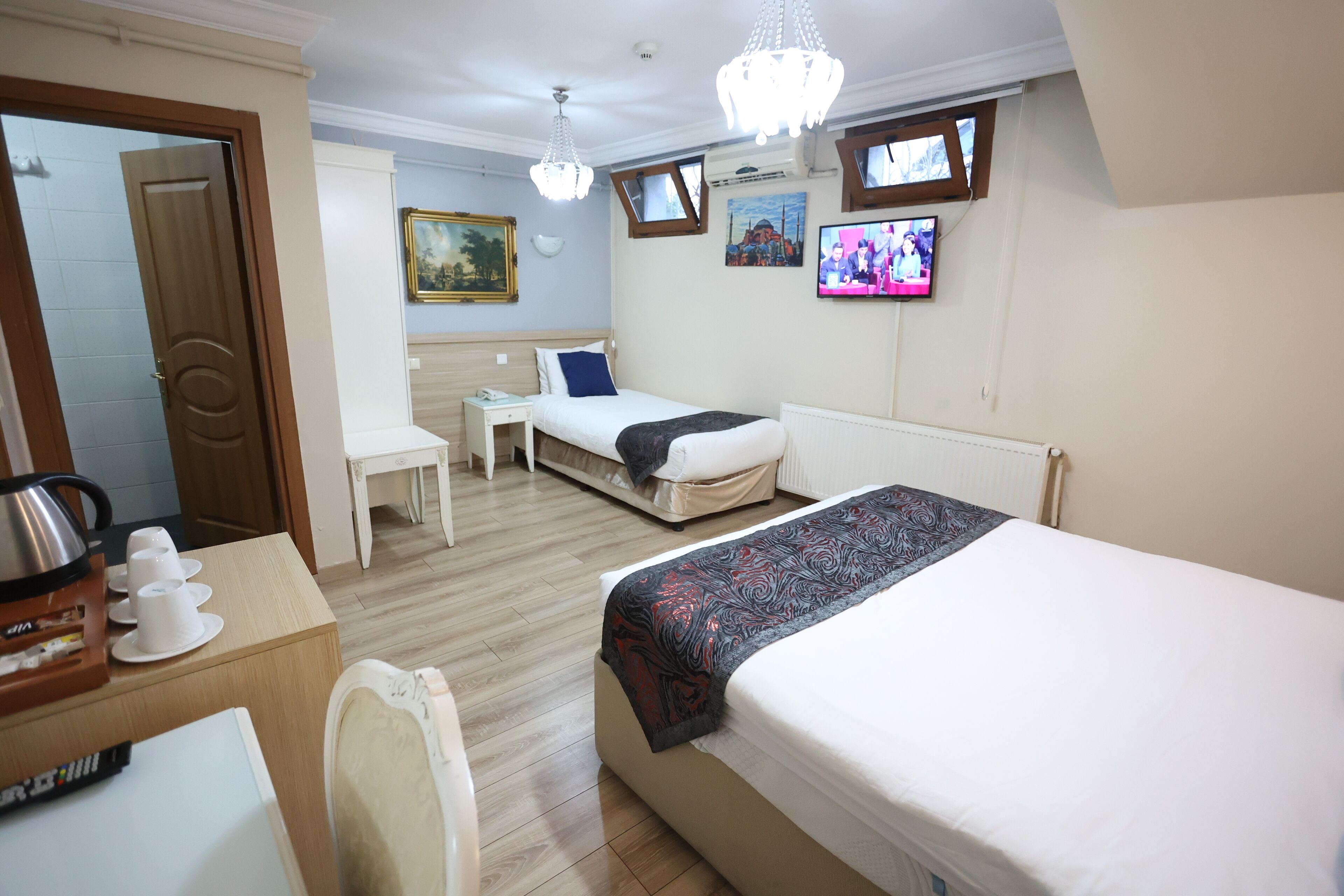 A hotel room with a bed, a desk with a kettle, a chair, a small table, and a television on the wall.