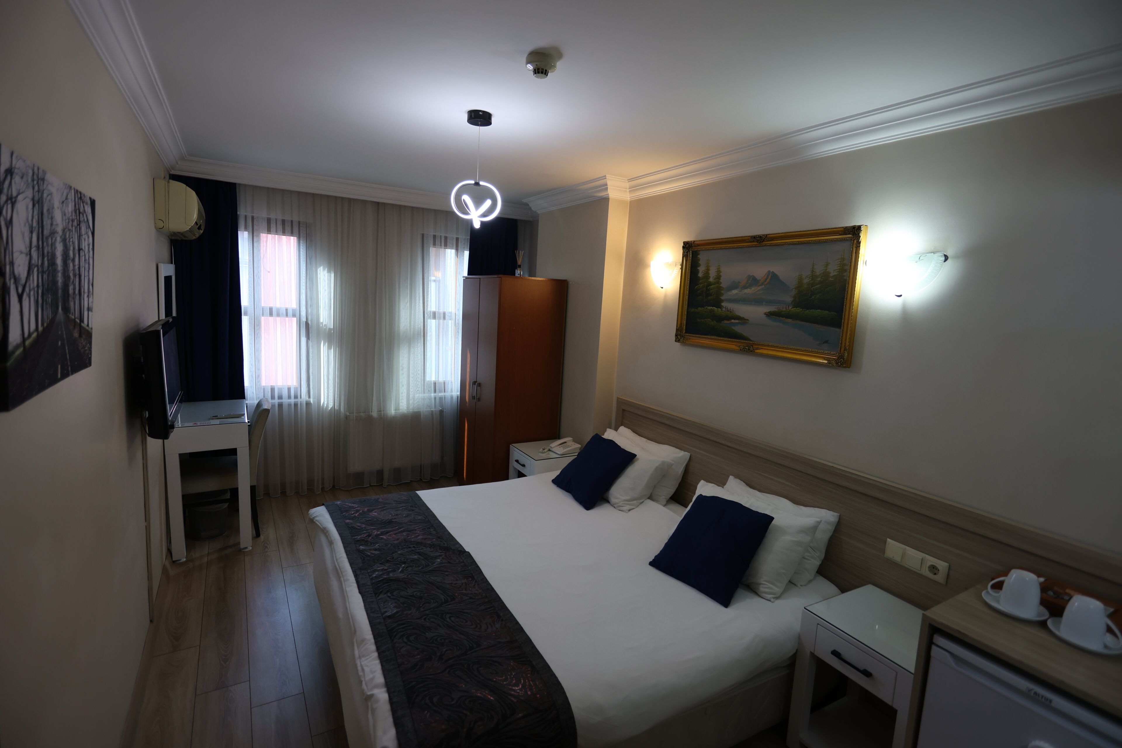 A hotel room with a bed, bedside tables, a desk, a wardrobe, and a painting on the wall.