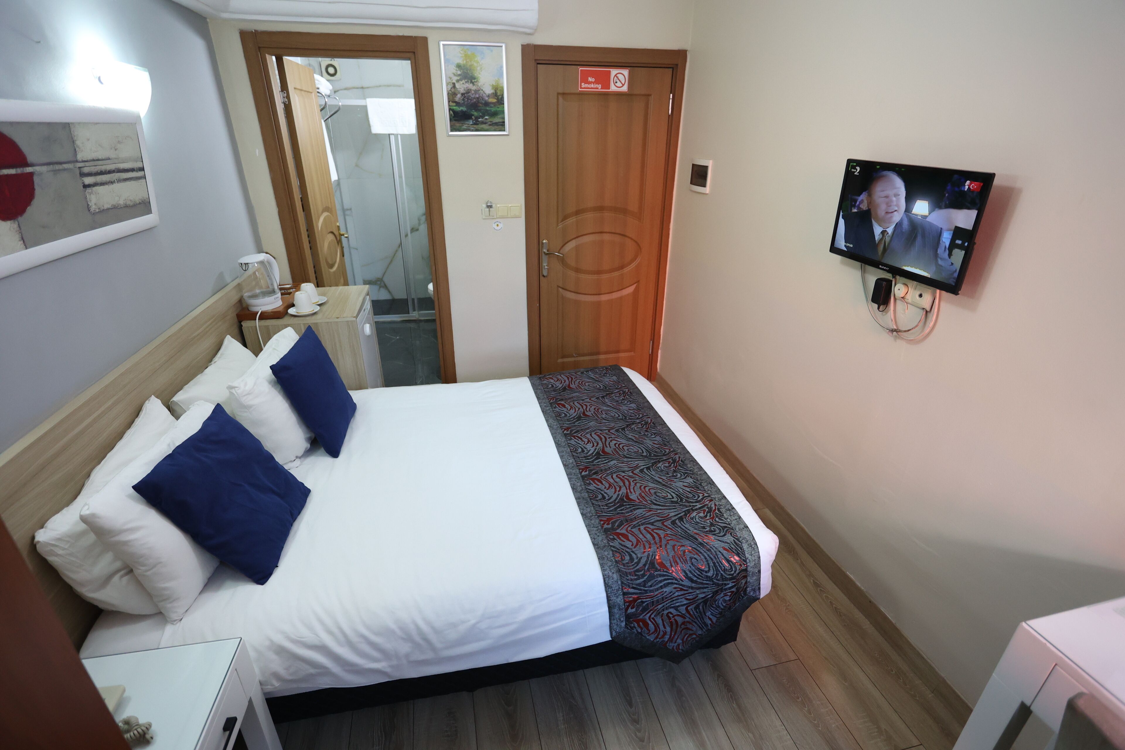 A hotel room with a bed, a television mounted on the wall, a door, and a small table with a phone and a cup.
