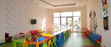 A brightly colored classroom with a long table and multiple chairs, a television on the wall, and a window providing a view of the outside.