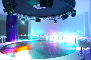 Un escenario con una plataforma circular, iluminación colgante y taburetes de bar dispuestos alrededor.