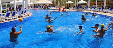 A swimming pool with people playing water polo, surrounded by lounge chairs and umbrellas.