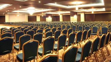 A large conference room with rows of blue chairs and tables, equipped with ceiling lights and a carpeted floor.
