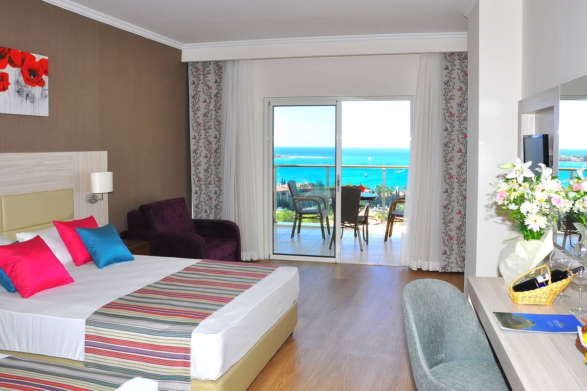 Habitación de hotel con cama, una silla, una mesa con un jarrón de flores y un balcón con vistas al mar.