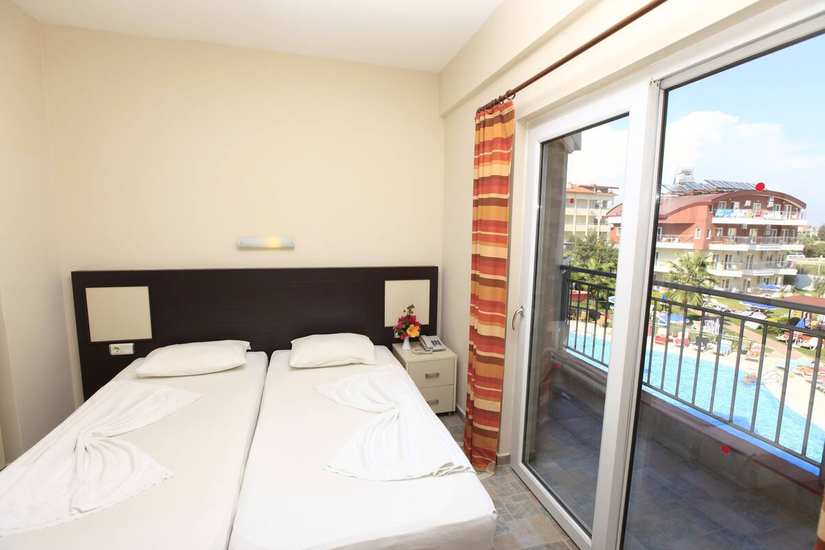Habitación de hotel con dos camas, una mesita de noche, una lámpara y vistas a una piscina y edificios.