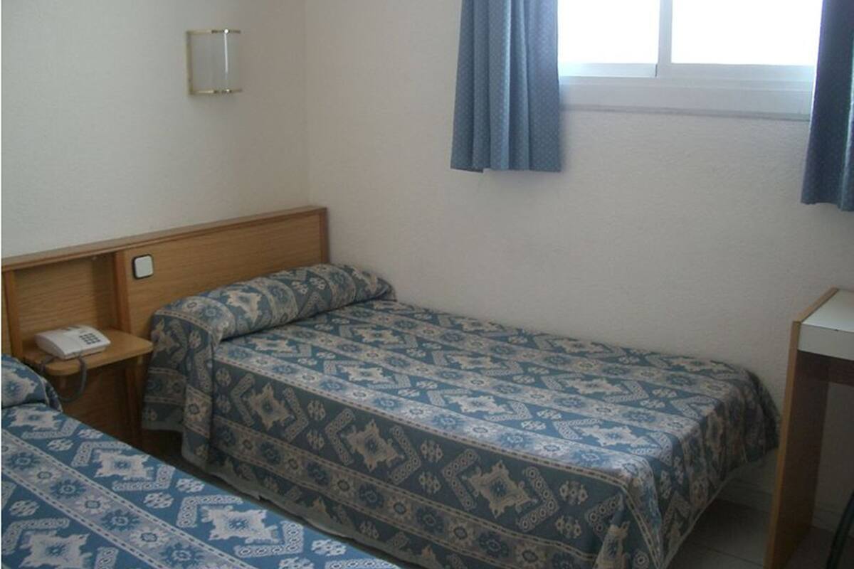 A small, simple room with two single beds, a bedside table with a phone, and a window with curtains.