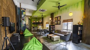 A modern living room with green bean bag chairs, a sofa, and a coffee table. There is a bookshelf and a dining area in the background. - PODs The Backpackers Home & Cafe (Kuala Lumpur)