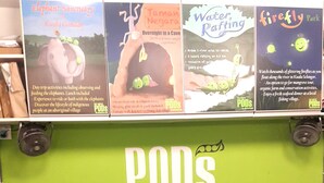 A reception desk with a green wall displaying various travel posters and a sign that reads "PODS the backpackers home the adventures hub". - PODs The Backpackers Home & Cafe (Kuala Lumpur)