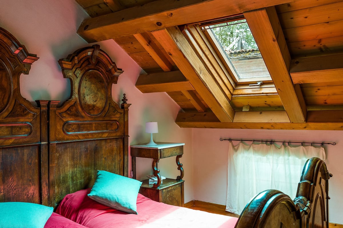 Een houten bed met schuine plafonds en een dakraam, een nachtkastje met lamp, en een raam met gordijnen.