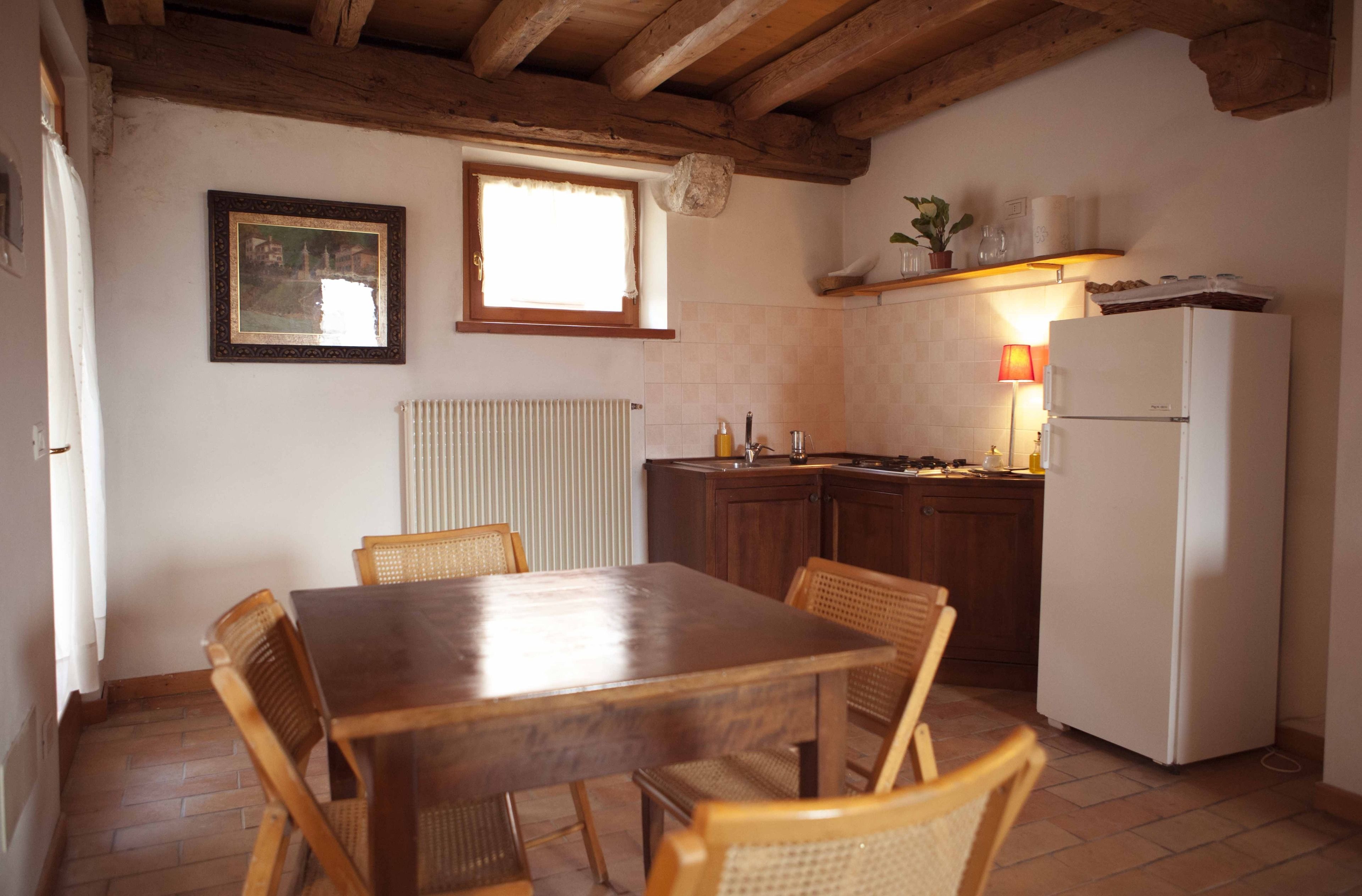 Uma sala de jantar com mesa e cadeiras de madeira, uma cozinha com geladeira e janela, e um radiador na parede.