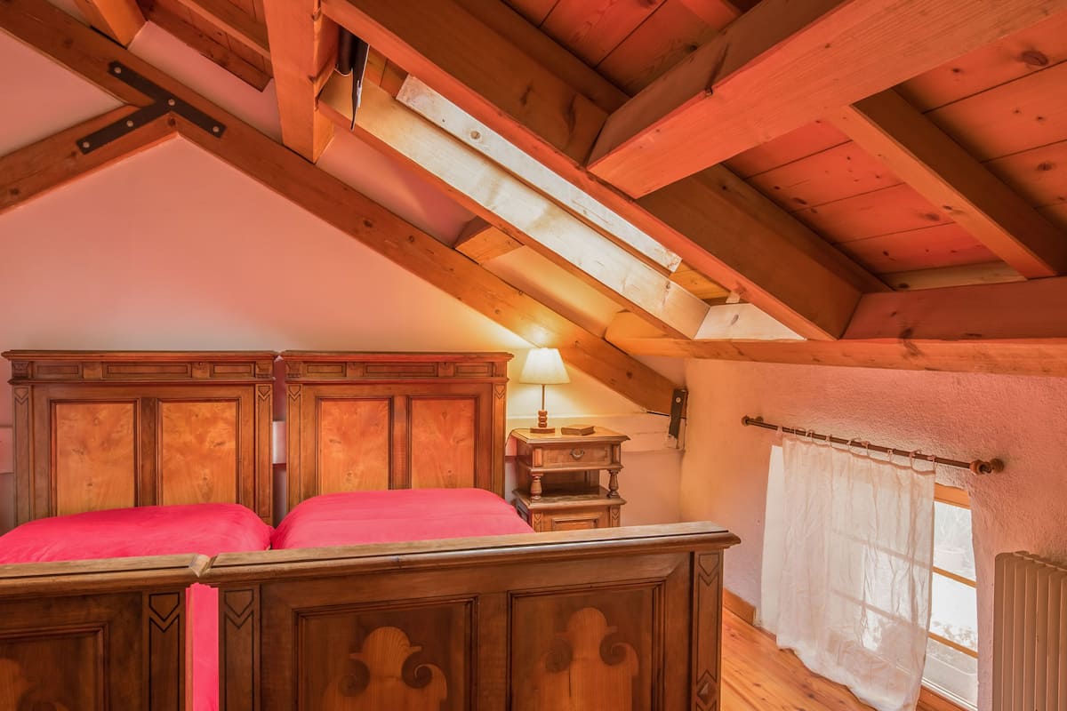Een slaapkamer met houten wanden en een schuine kap, een bed met rode beddengoed, een nachtkastje, een lamp en een raam met gordijnen.