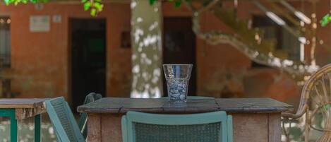 A rustic outdoor seating area with a table and chairs, a glass vase with stones, and a tree providing shade.
