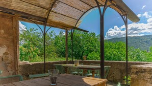 Une terrasse extérieure avec des tables et des chaises en bois, un mur de pierre et une vue sur des arbres et des collines verdoyantes.