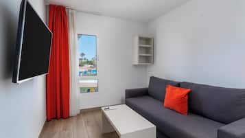 Un salon moderne avec un canapé gris, un coussin rouge, une table basse blanche, un téléviseur fixé au mur et une fenêtre avec des rideaux.