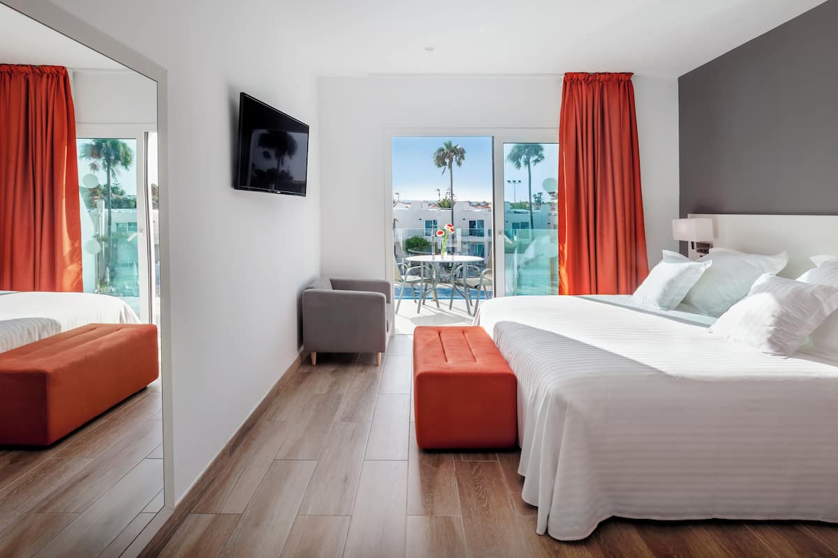 Habitación de hotel moderna con una cama grande, zona de estar y balcón con vistas a palmeras y edificios.