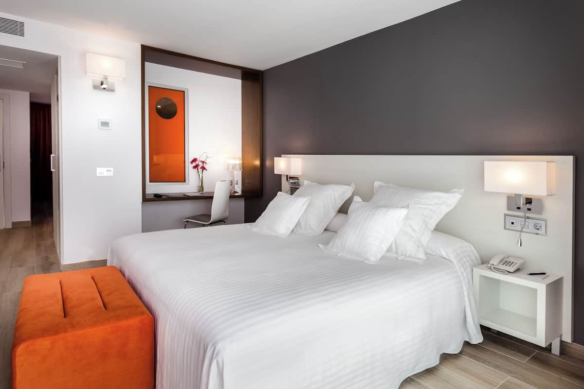 Camera d'albergo moderna con un letto grande, due comodini con lampade, un poggiapiedi arancione e uno specchio con cornice rossa.