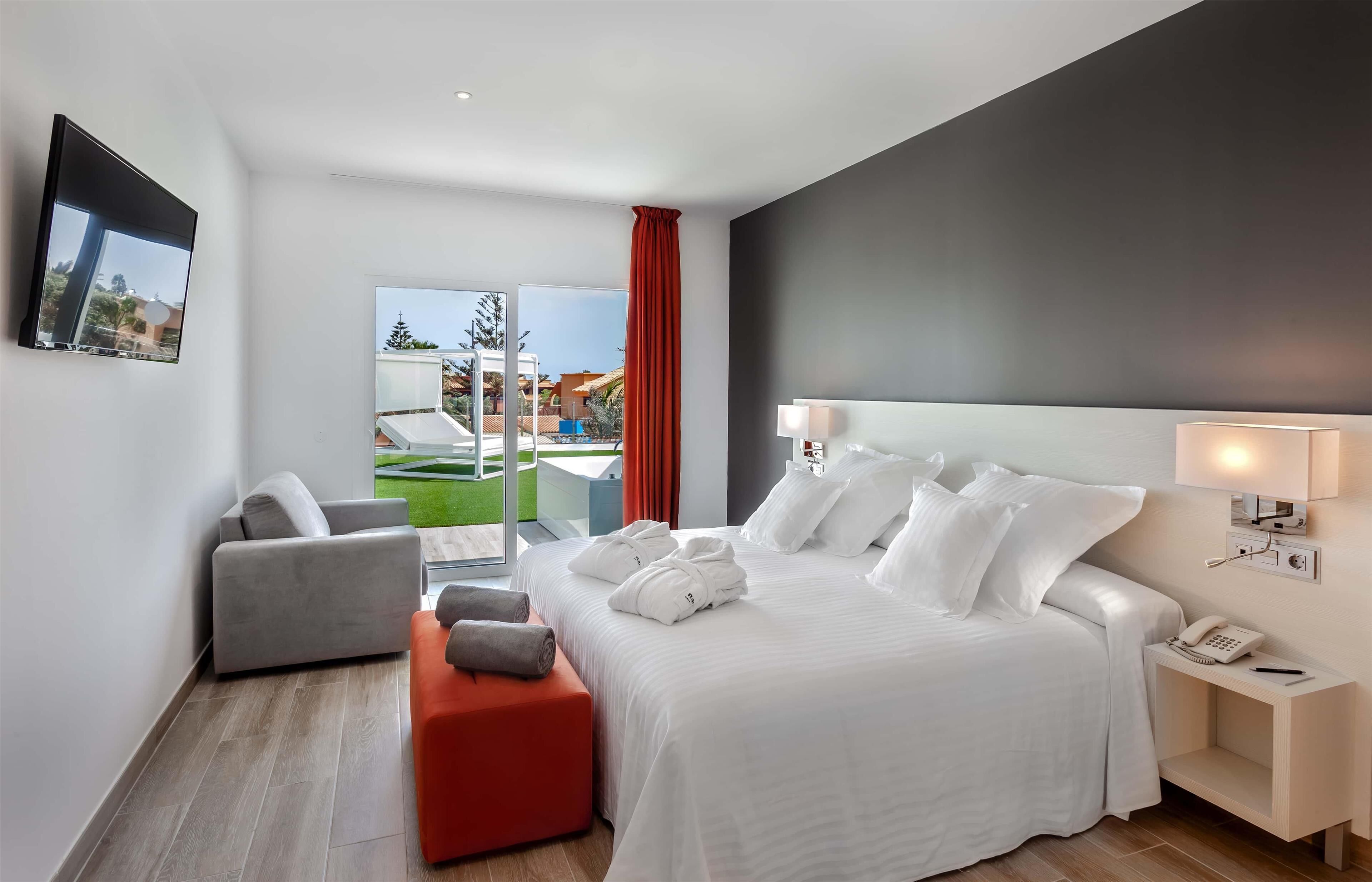 A hotel room with a bed, a chair, a small table, a phone, and a window with a view of a pool and greenery.