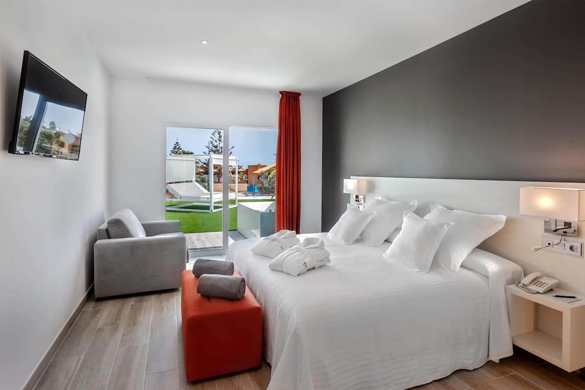 A hotel room with a bed, a chair, a small table, a phone, and a window with a view of a pool and greenery.