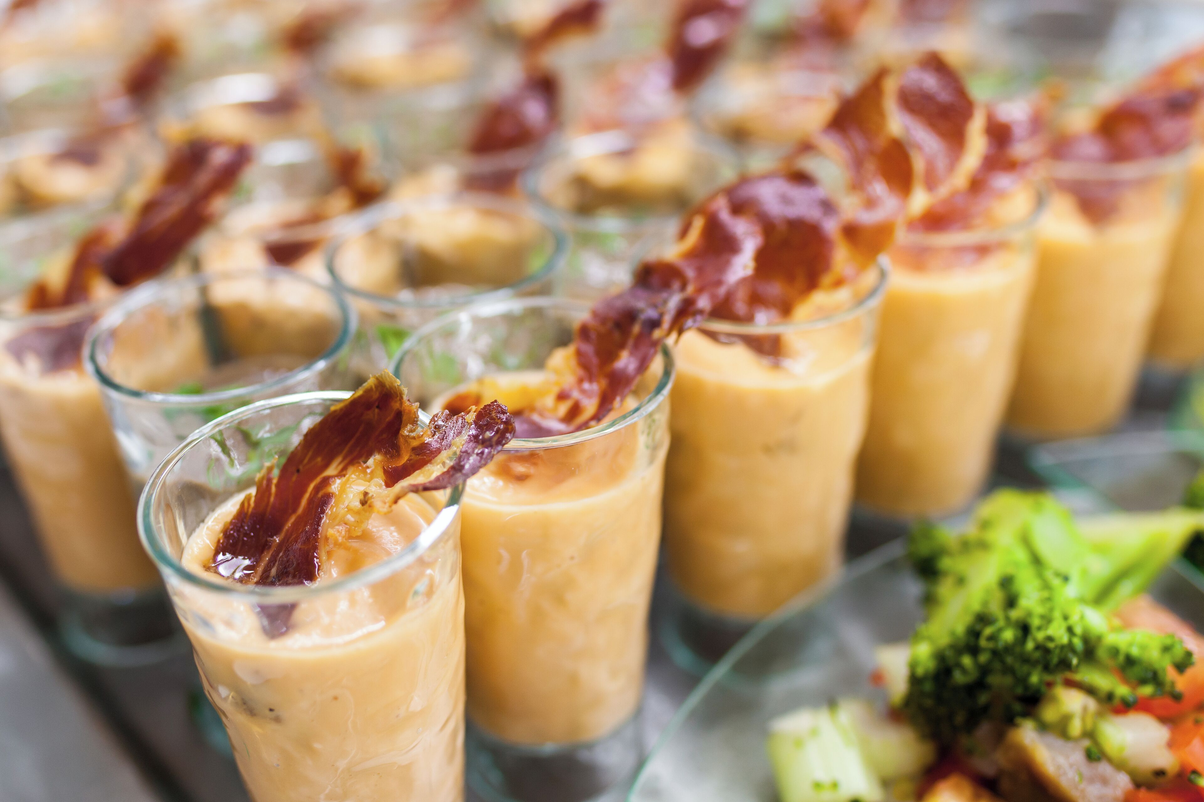 A row of glasses filled with a creamy beverage, garnished with crispy bacon strips, and a plate of mixed vegetables in the background.