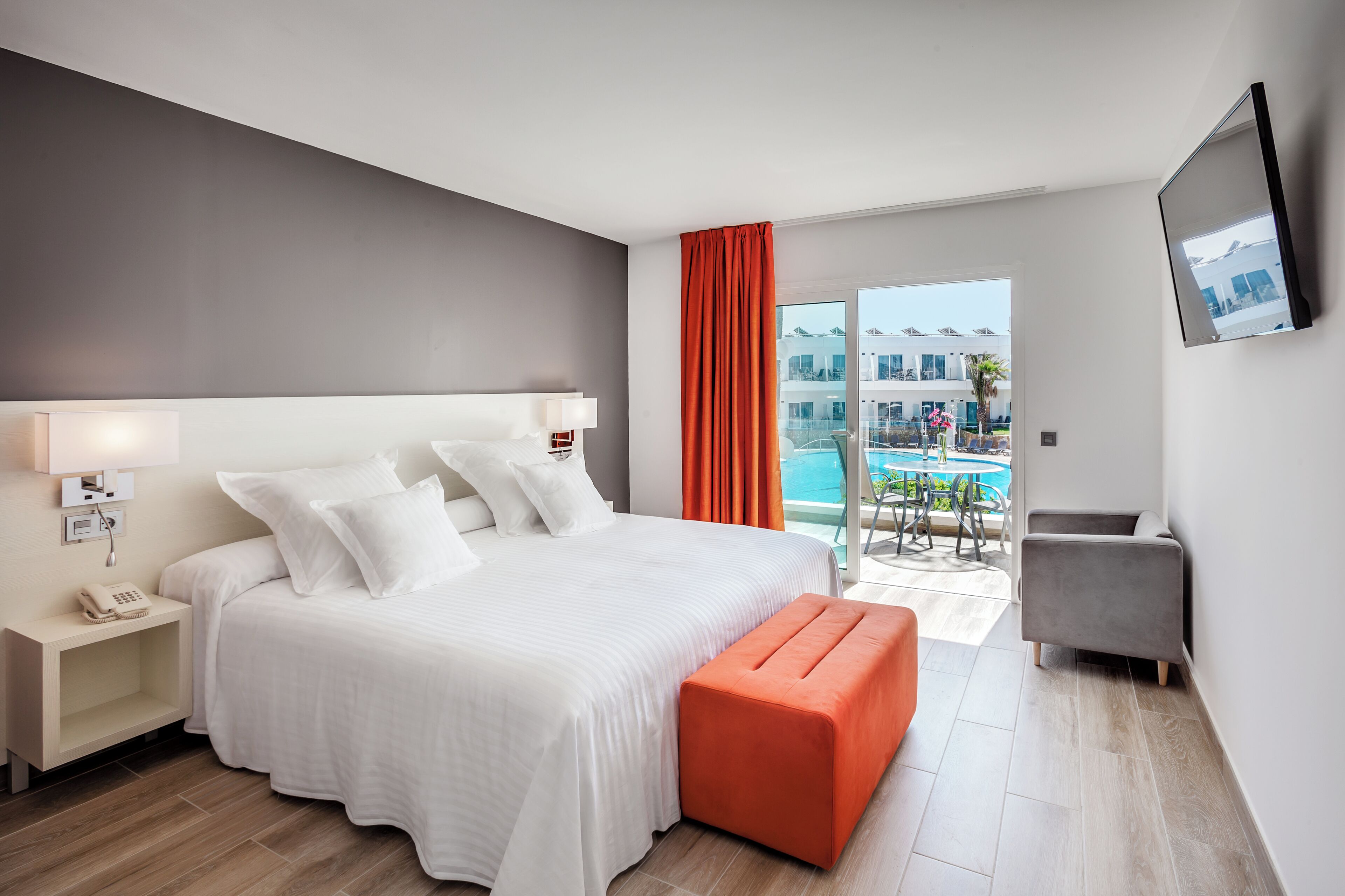 A modern hotel room with a large bed, a bedside table with a phone, a bright orange ottoman, a gray armchair, and a balcony with a pool view.