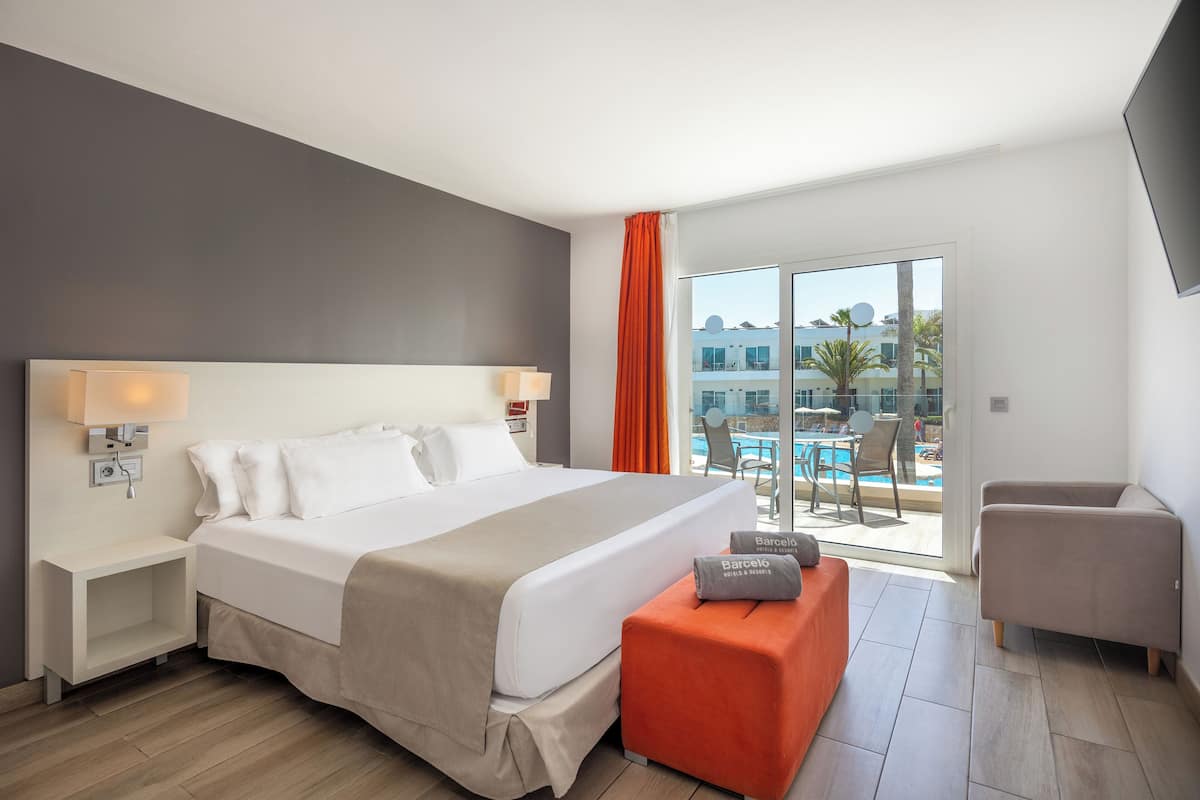 Camera d'albergo moderna con un ampio letto, un comodino, un poggiapiedi arancione e vista su una piscina.