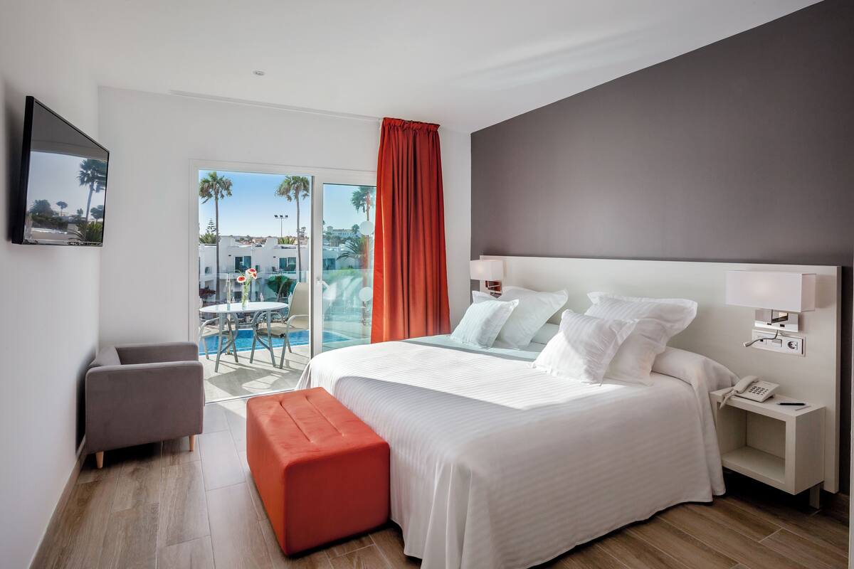 Una camera d'albergo moderna con un letto grande, una scrivania, una sedia e vista su una piscina e palme.