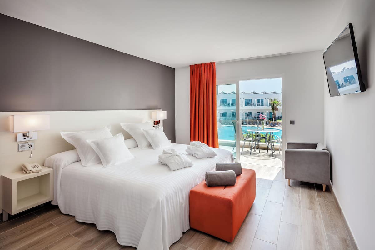 Una camera d'albergo moderna con un letto grande, un poggiapiedi arancione, una poltrona grigia e un balcone vista piscina.