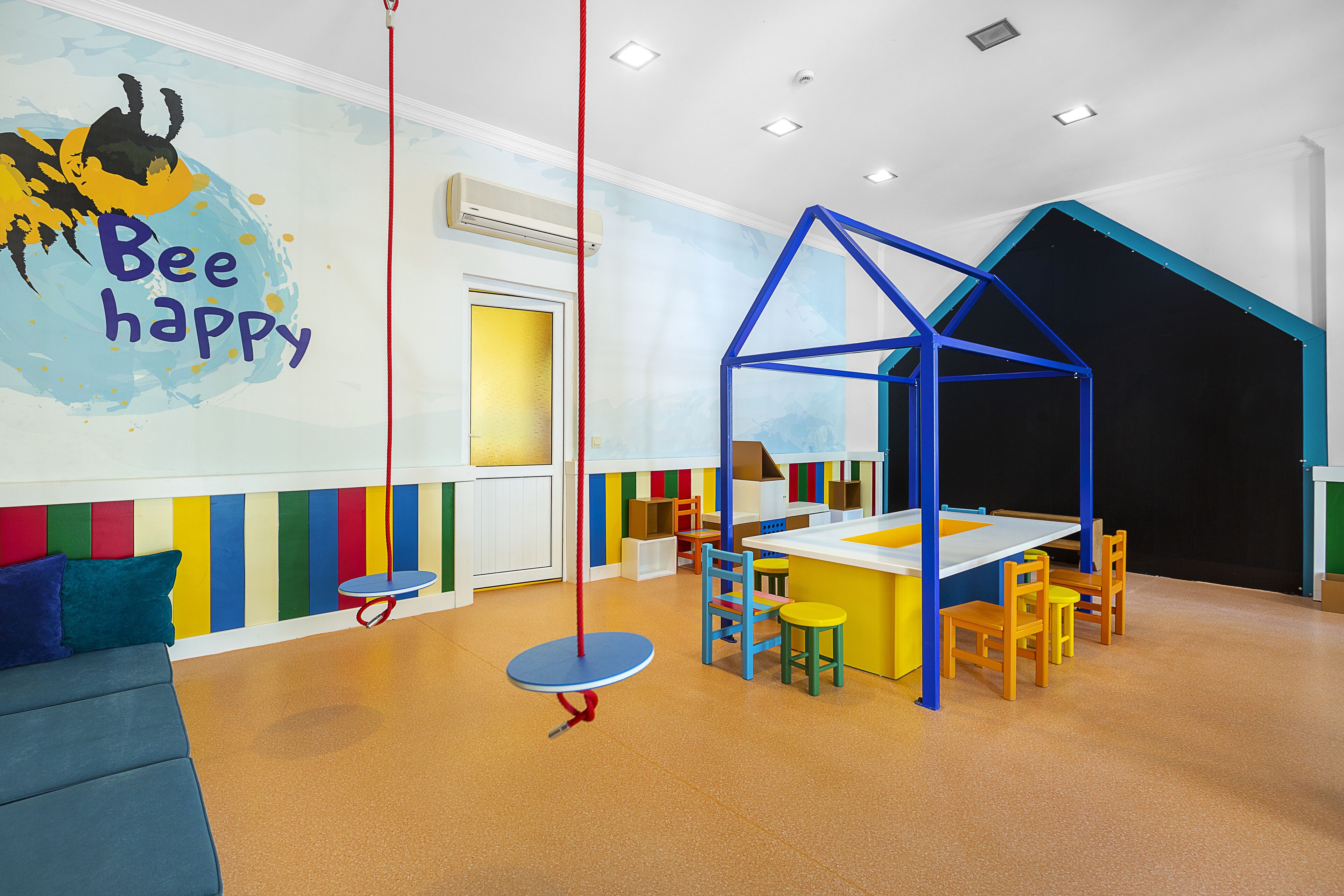 A children's playroom with a colorful wall mural, a blue playhouse, and a seating area with a sofa and chairs.