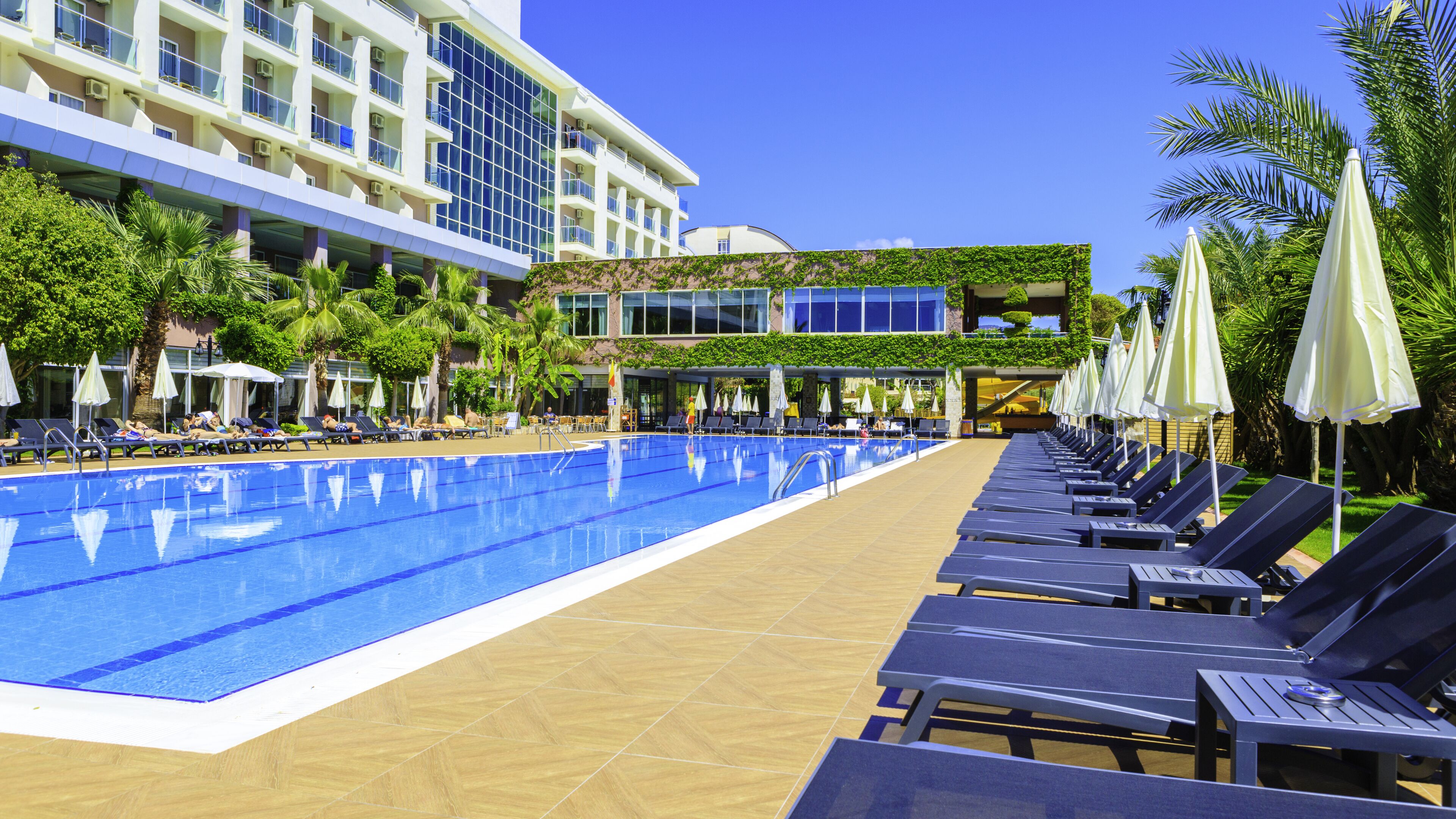 A pool area with lounge chairs and umbrellas, surrounded by greenery and a building with multiple floors and balconies.