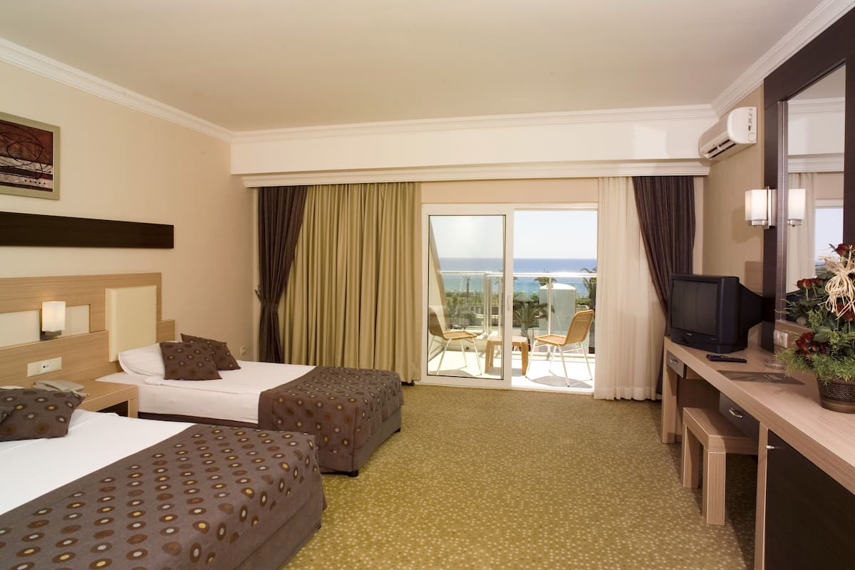A hotel room with two beds, a desk with a television, and a balcony with a view of the sea.