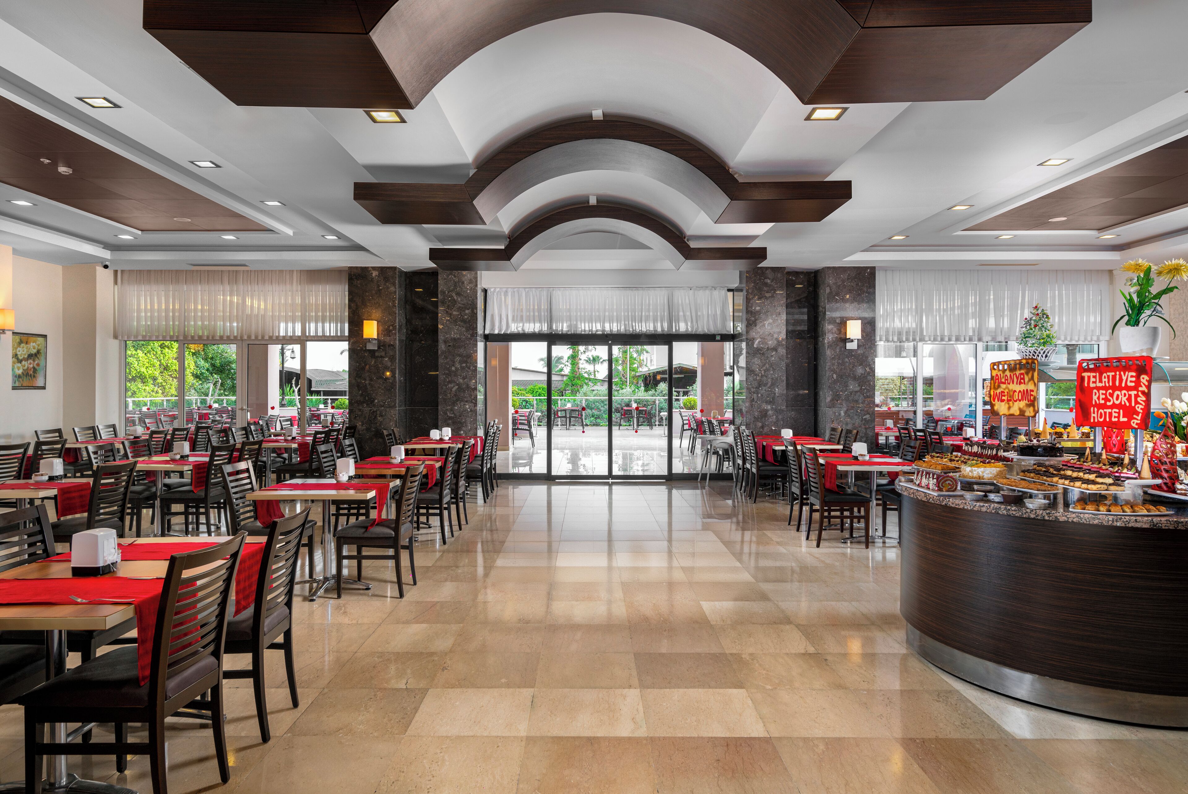 A spacious hotel dining area with multiple tables and chairs, a central reception desk, and large windows offering a view of the outdoors.